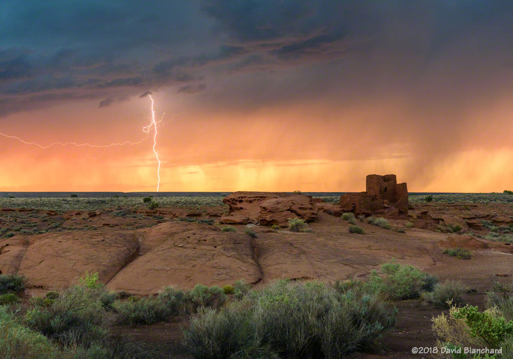 The setting sun lights up rain curtains as a brilliant lightning bolt strikes behind Wukoki Pueblo in Wupatki National Monument.