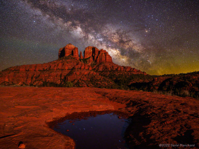The Milky Way is lower in the sky and is combined with a foreground image containing star reflections in the small pool of water.